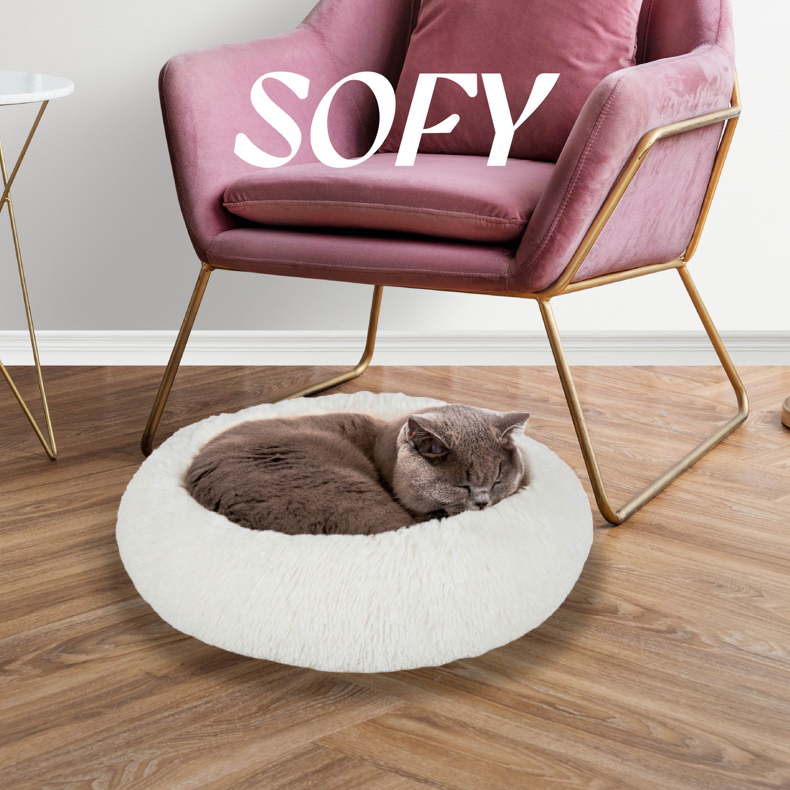 Gray cat sleeping in round white plush pet bed on wooden floor beside pink velvet chair