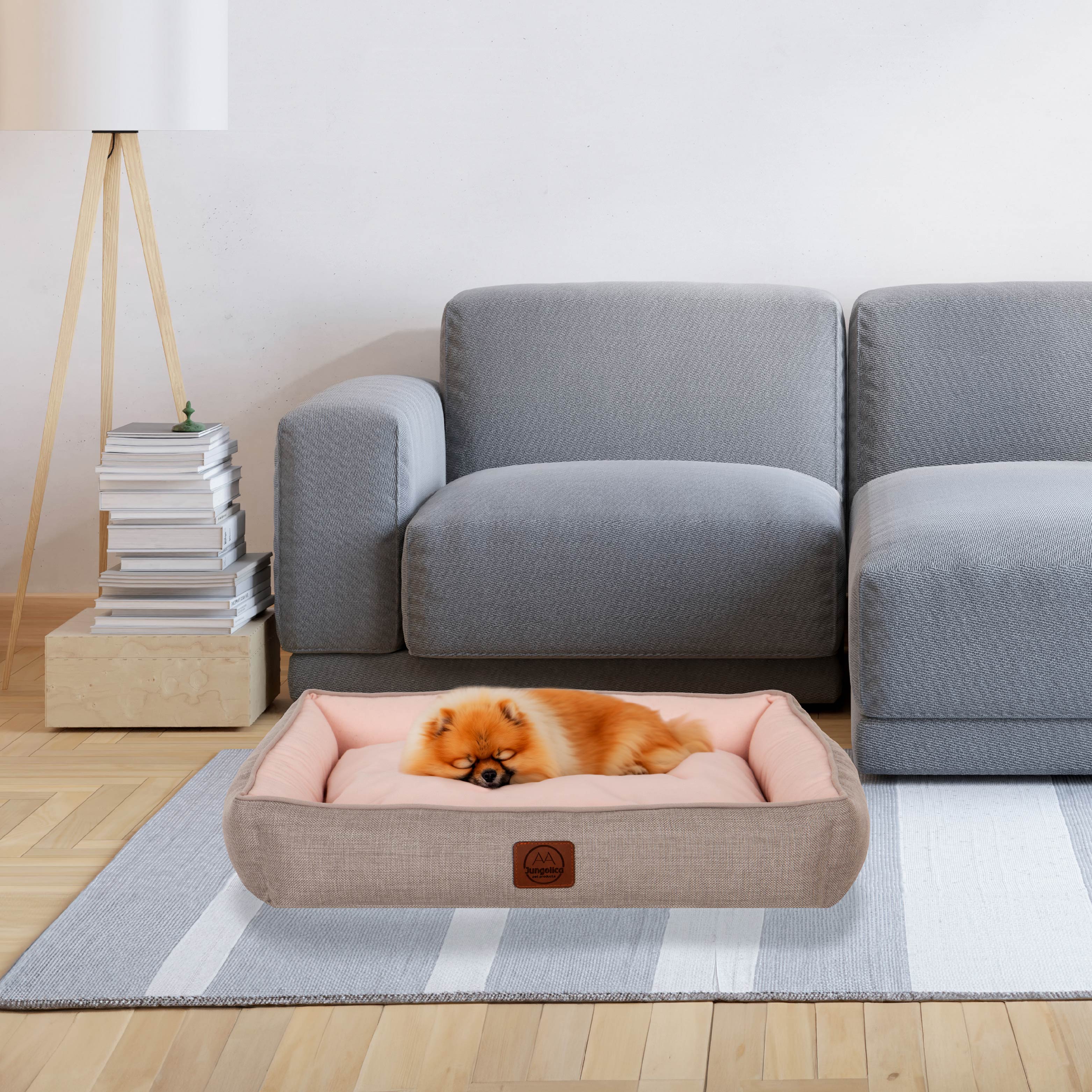 Small fluffy dog sleeping in a pink pet bed on a striped rug beside a grey sofa